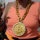 A person in a salmon-colored shirt wears a massive fake gold chain around their neck the the Bitcoin symbol at the end of it.